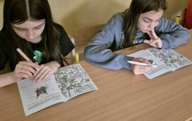 Dwie dziewczynki siedzą w ławce szkolnej, rozwiązują zadania w książce.