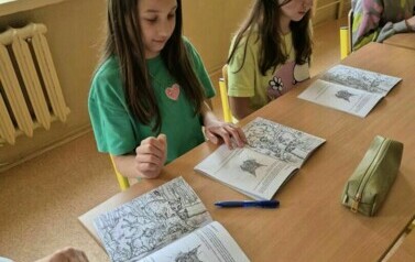 Dwie dziewczynki siedzą w ławce szkolnej, rozwiązują zadania w książce