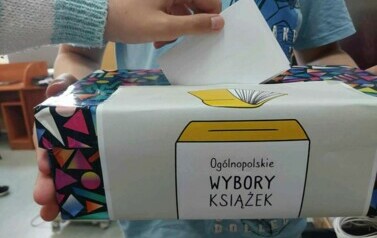 Wyciągnięta ręka w jasnym swetrze wrzuca kartkę do kolorowego pudełka z napisem Og&oacute;lnopolskie Wybory Książek kt&oacute;re trzyma dziecko w niebieskiej bluzce.