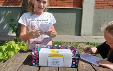 Jasny słoneczny dzień Dziewczynka w jasnej koszulce wrzuca kartkę do kolorowego pudelka z napisem Og&oacute;lnopolskie Wybory Książek W tle ściana z czerwonych cegieł.