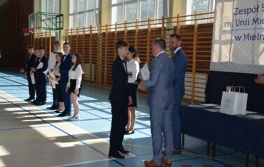 Uczeń klasy &oacute;smej odbiera świadectwo ukończenia szkoły podstawowej od dyrektora oraz wychowawcy podczas uroczystego zakończenia roku szkolnego.