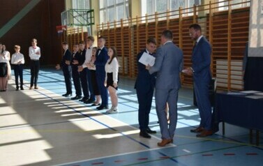 Uczeń klasy &oacute;smej odbiera świadectwo ukończenia szkoły podstawowej od dyrektora plac&oacute;wki podczas uroczystego zakończenia roku szkolnego.