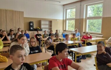 Grupa uczni&oacute;w w sali lekcyjnej siedzi w ławkach szkolnych.