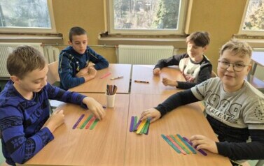 Chłopcy siedzą przy stolikach, na kt&oacute;rych lezą kolorowe patyczki z tabliczką mnożenia.