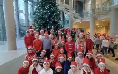 Trzydzieścioro dzieci w mikołajowych czapeczkach pozuje przed wielką choinką na korytarzu opery.