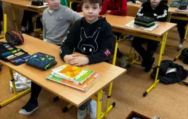 Uczniowie siedzą w ławkach szkolnych, na podłodze obok ławki leży szuflada z książkami