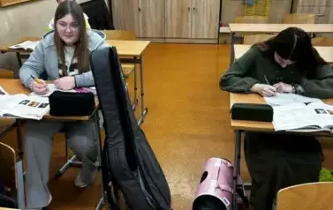 Uczennice siedzą w ławkach szkolnych obok kt&oacute;rych stoją futerały z książkami