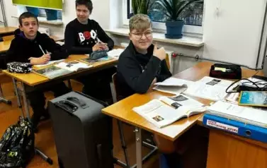 Chłopcy siedzą w ławkach szkolnych. Obok stoi walizka.