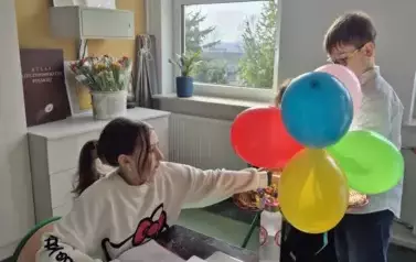 Chłopiec częstuje cukierkami dziewczynkę, kt&oacute;ra siedzi przy stoliku, na kt&oacute;rym znajdują się książki i zeszyty
