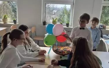 Trzy dziewczynki siedzą przy stoliku, a przy nich stoi chłopiec w białej koszuli z czarną muchą