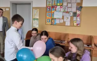 Chłopiec z wiklinowym koszem ozdobionym kolorowymi balonami częstuje uczennice klasy &oacute;smej cukierkami