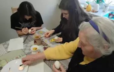 Uczennice z seniorką siedzące przy stole i malujące jajka