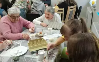 Uczennice i seniorzy siedzący przy stole malujący jajka.