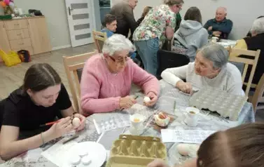 Uczniowie z seniorami siedzą przy stole i malują jajka.