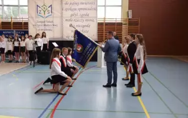 Uczniowie w galowych strojach z przewieszonymi biało-czerwonymi szarfami  klęczą przed sztandarem szkolnym trzymanym przez dyrektora szkoły.