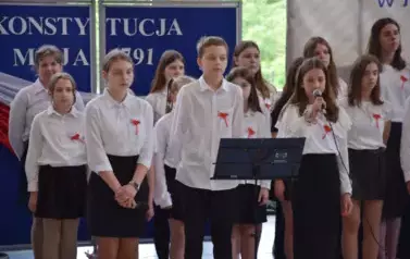 Uczniowie w czarno-białych strojach śpiewją podczas uroczystości.