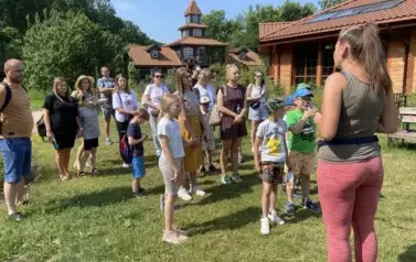 Dzieci z rodzicami stoją na trawie i słuchają co m&oacute;wi pani