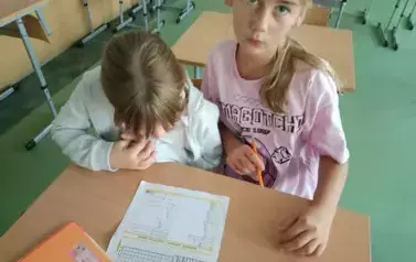 Uczennice siedzą w ławce i wykonują karty pracy