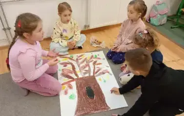 Pięcioro dzieci siedzi na szarym dywanie dookoła plakatu z drzewem
