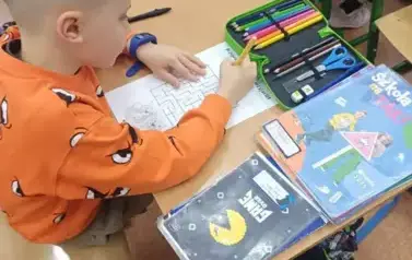 Chłopiec siedzi przy ławce szkolnej, na kt&oacute;rej leżą książki, pi&oacute;rniki z kolorowymi kredkami i przyboram