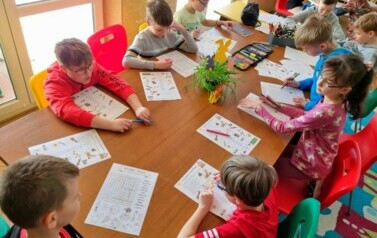 Grupa dzieci siedzi dookoła stołu, rozwiązują łamigł&oacute;wki. Na stole znajdują się kartki, mazaki.
