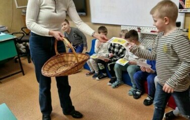 Kobieta w beżowej bluzce i granatowych spodniach  trzyma koszyk i wręcza upominek chłopcu 