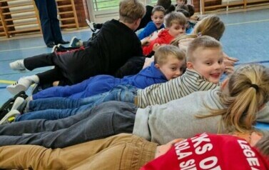 Uczniowie leżą na brzuchach obok siebie, za nimi stoi nauczycielka