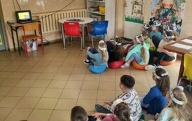 Dzieci siedzą na dywanie i ogladają animacje wyświeloną na projektorze, dotyczącą segregacji odpad&oacute;w.