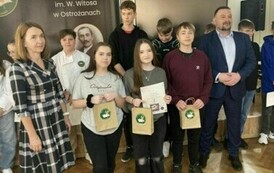 Grupa uczni&oacute;w z dyplomami i papierowymi torbami. Wśr&oacute;d nich kobiata i mężczyzna ubrani w eleganckie stroje.