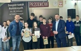 Grupa uczni&oacute;w pozuje do zdjęcia trzymając dyplomy oraz papierowe torby. Wśr&oacute;d nich dw&oacute;ch mężczyzn. Jeden z nich ubrany jest w garnitur, drugi w str&oacute;j jeansowy.