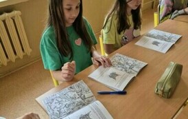 Dwie dziewczynki siedzą w ławce szkolnej, rozwiązują zadania w książce