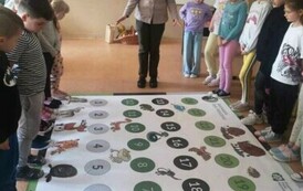Uczniowie ustawieni w dw&oacute;ch rzędach, pomiędzy nimi na podłodze rozłożona gra planszowa. Przed dziećmi stoi kobieta.
