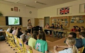 Uczniowie w sali lekcyjnej, siedzą w ławkach szkolnych ustawionych w kształcie podkowy. Przed nimi stoi kobieta. W tle, na ścianie widać monitor interaktywny oraz tablicę szkolną.