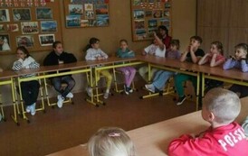Uczniowie w sali lekcyjnej, siedzą w ławkach szkolnych ustawionych w kształcie podkowy