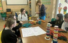 Uczennice siedzące dookoła stołu, obok kobieta. Na stole białe kartki, stojaki z bransoletkami.