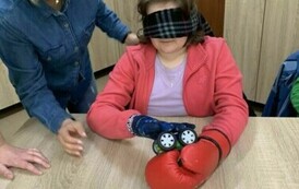 Dziewczynka w czerwonej bluzie, fioletowej koszulce, z zasłoniętymi oczami, w rękawicach na dłoniach, siedzi przy stole i trzyma samochodzik-zabawkę