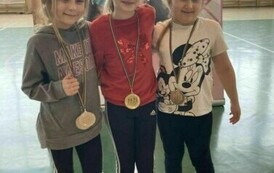 Trzy dziewczynki w strojach sportowych, stojące obok siebie, każda z nich ma medal przewieszony na szyi (złoto, srebro, brąz).