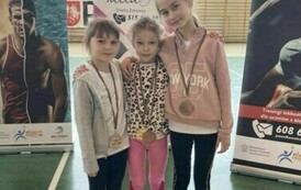 Trzy dziewczynki w strojach sportowych, stojące obok siebie, każda z nich ma medal przewieszony na szyi