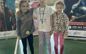 Trzy dziewczynki w strojach sportowych, stojące obok siebie, każda z nich ma medal przewieszony na szyi.