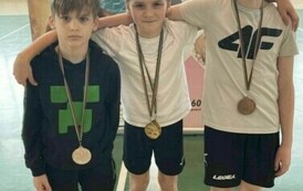 Trzech chłopc&oacute;w w strojach sportowych, stojących obok siebie, każdy z nich ma medal przewieszony na szyi.