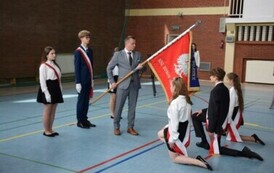 Tr&oacute;jka uczni&oacute;w w galowych strojach z przewieszonymi biało-czerwonymi klęczy przed sztandarem szkolnym trzymanym przez dyrektora plac&oacute;wki