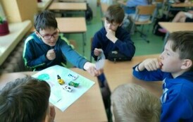 Pięciu chłopc&oacute;w siedzi przy stoliku szkolnym. Na blacie stołu znajduje się kartka i małe samochodziki.