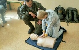 Chłopiec wykonuje reanimacje na leżącym na podłodze fantomie. Obok chłopca znajduje się mężczyzna w wojskowym mundurze.