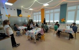 Dzieci siedzą w jasnej sali, przy stolikach, na kt&oacute;rych znajdują się kolorowe obrusy