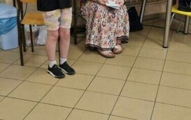 Kobieta siedzi na krześle. Obok niej stoi dziewczyna, trzyma przed sobą kartkę.