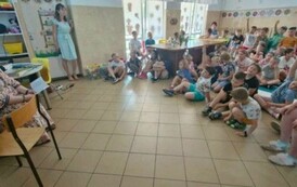 Uczniowie siedzą naprzeciw kobiety czytającej książkę. Kilkoro z nich unosi rękę.