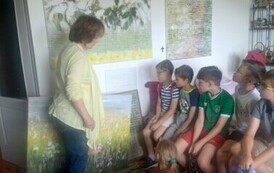 Pani malarka stoi przy ścianie, na kt&oacute;rej wiszą duże obrazy przedstawiające kwiaty i opowiada o nich dzieciom siedzącym w pobliżu.