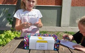 Jasny słoneczny dzień Dziewczynka w jasnej koszulce wrzuca kartkę do kolorowego pudelka z napisem Og&oacute;lnopolskie Wybory Książek W tle ściana z czerwonych cegieł.