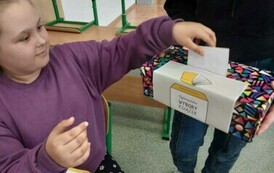 Dziewczynka wrzuca kartkę do kolorowego pudełka z napisem Og&oacute;lnopolskie Wybory Książek, kt&oacute;re tzryma inna osoba. W tle ławka szkolna.