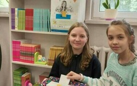 Dwie dziewczynki w szkolnej bibliotece biorą udział w wyborach książek. Jedna w czarnej bluzie trzyma pudełko druga wrzuca kartkę do głosowania. W tle regał z książkami i maskotkami
